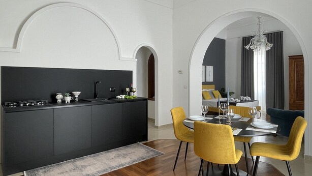 A modern kitchen with a black countertop and white walls, leading to a dining area with yellow chairs and a chandelier. - Ortigia Suite 1939 (Siracusa)