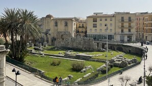 A historical site with ancient ruins, a green lawn, and modern buildings in the background. - Ortigia Suite 1939 (Siracusa)