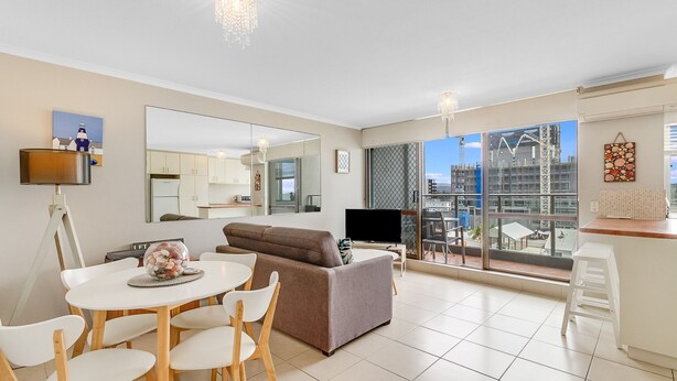 Een moderne woonkamer met een bank, eettafel en stoelen. Een televisie is aan de muur bevestigd, en er is een balkon met uitzicht op de stad.