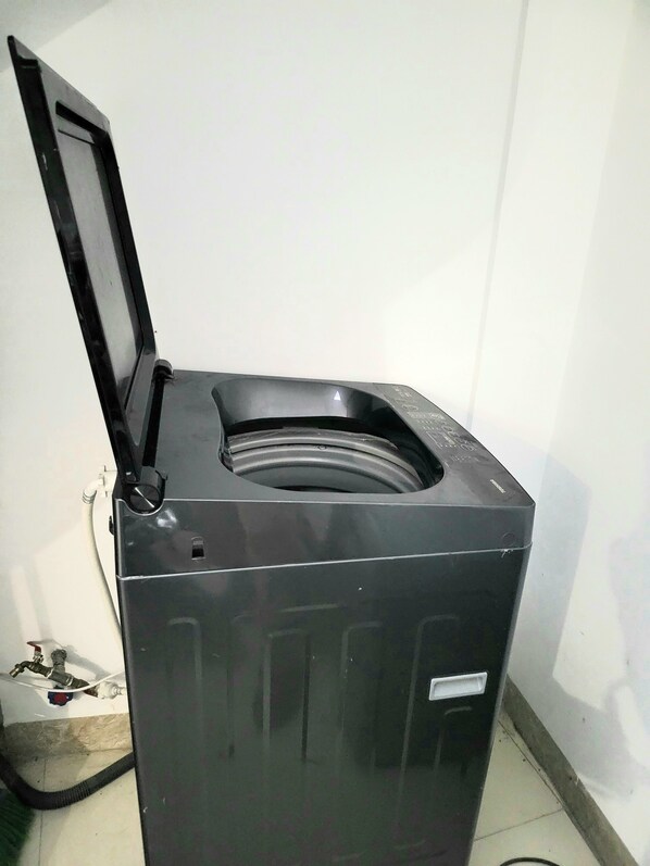 A compact, black, wall-mounted laundry machine with a transparent door, a control panel on the front, and a hose connected to the side.