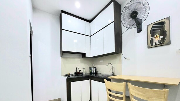 A compact kitchen with white cabinets, a black countertop, a sink, and a fan. There are two wooden chairs and a small table. - Ruby Relax Home (Hanoi)