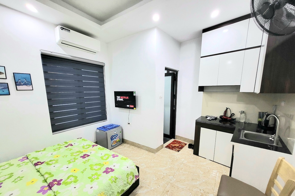 A compact room with a bed, a small kitchenette, a TV, and a window with blinds.