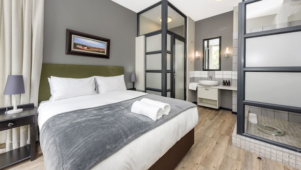 Een moderne hotelkamer met een groot bed, een nachtkastje met lamp, een badkamer met wastafel en spiegel, en een ingelijst schilderij aan de muur.
