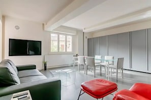 A modern living room with a grey sofa, red ottoman, and a flat-screen TV mounted on the wall. - pieno centro storico a poche centinaia di metri dal mare, appartamento di 95 mq (Pesaro)