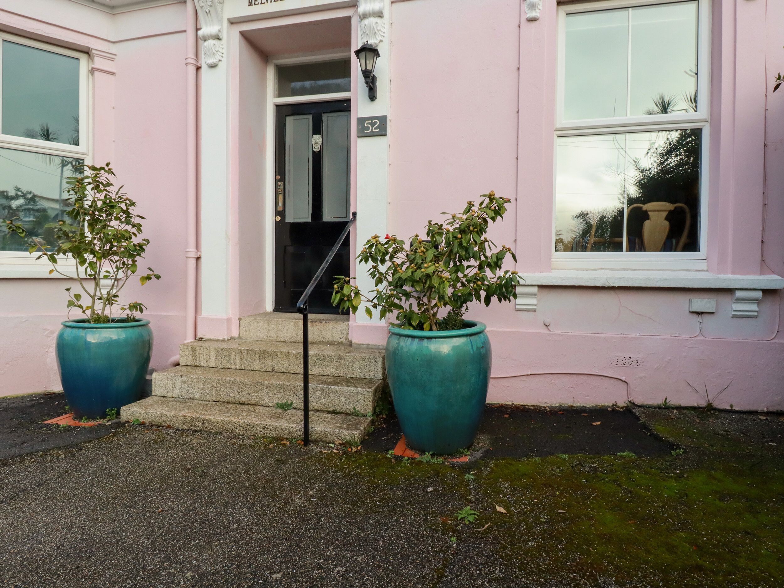 A building with a pink facade, a black door, and the number 52 on the door. There are two large blue planters on the ground in front of the building.