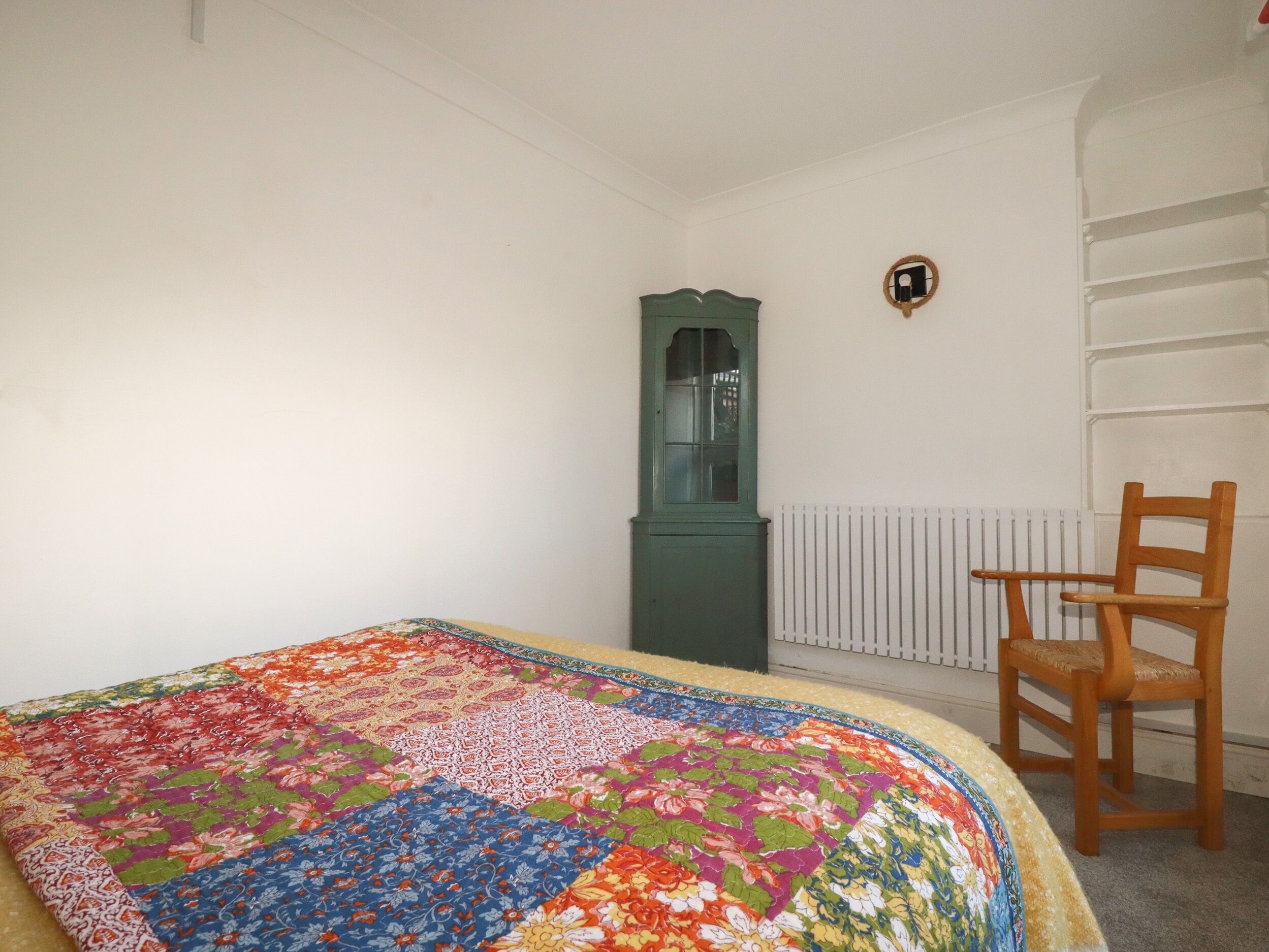 A bedroom with a colorful quilt, a wooden chair, a green cabinet, and a radiator.