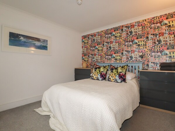 Une chambre à coucher comprenant un lit, une commode, un mur orné d’un motif coloré représentant des bâtiments, et un tableau encadré représentant un paysage marin.