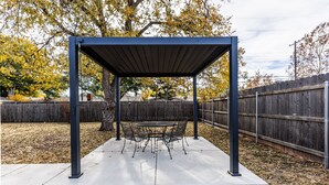 A covered patio area with a table and chairs, surrounded by a wooden fence and trees. - Walk to Groceries—Sleeps 8—Game Room + Patio + Parking for 4 (Lubbock)