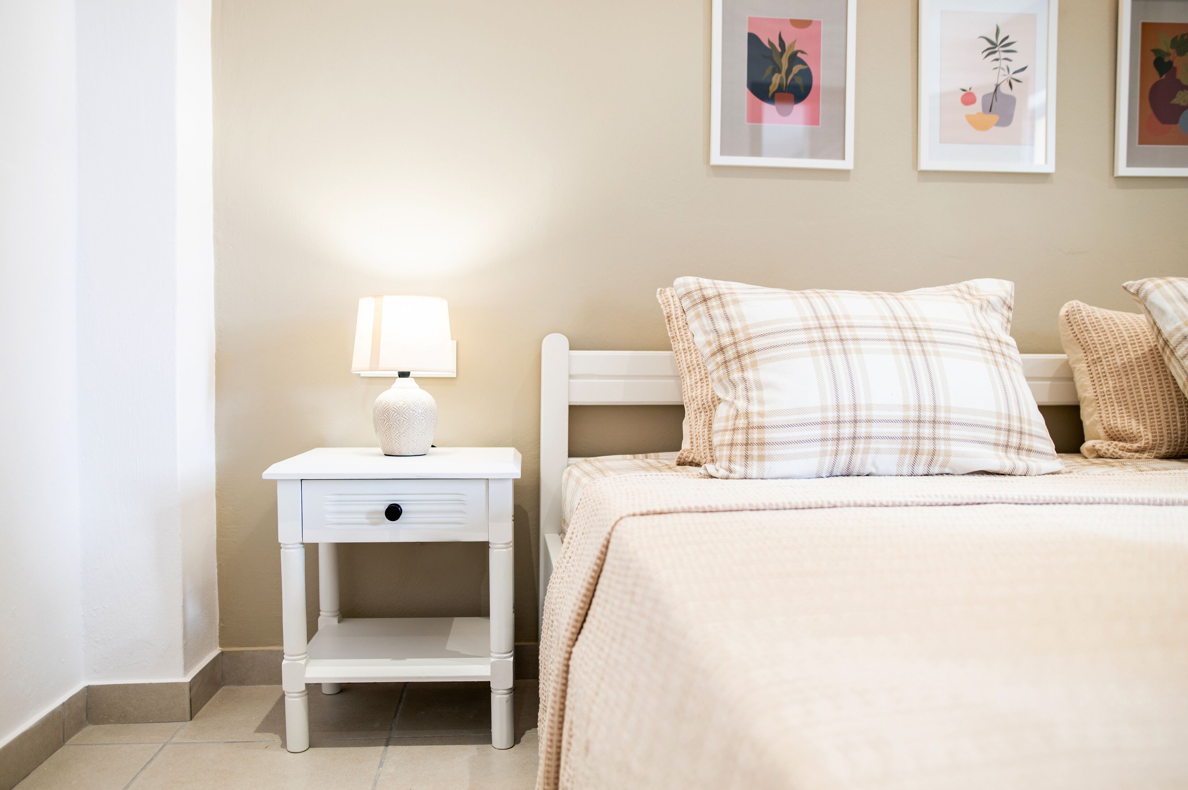 A neatly arranged bedroom with a white nightstand, a lamp, a bed with plaid bedding, and three framed pictures on the wall.