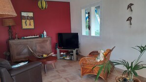 Uma sala de estar com um sofá, uma poltrona de vime, uma televisão, um armário e um vaso de plantas.
