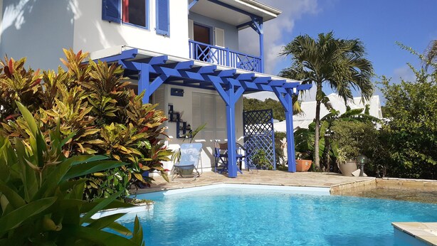 Uma área de piscina com uma pérgula azul, um edifício branco com venezianas azuis e plantas tropicais.