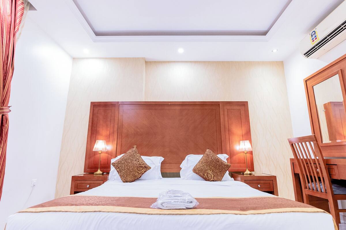 A hotel room with a large bed, two bedside lamps, a wooden headboard, a mirror, and a wooden chair.