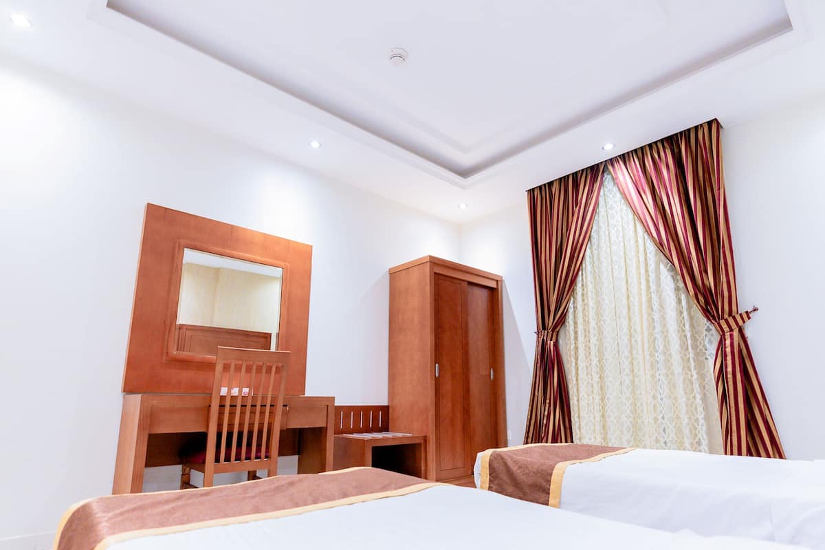 A hotel room with two beds, a wooden desk, a chair, a wardrobe, and a large mirror on the wall.