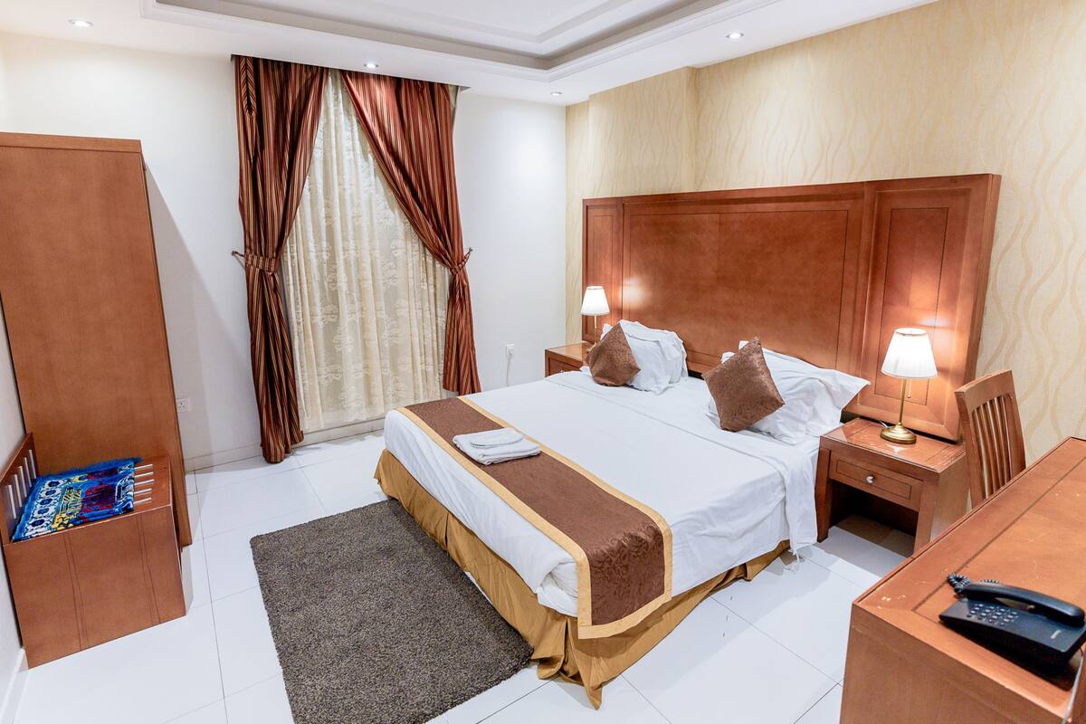 A hotel room with a large bed, two bedside tables with lamps, a wooden headboard, a wardrobe, and a desk with a telephone.