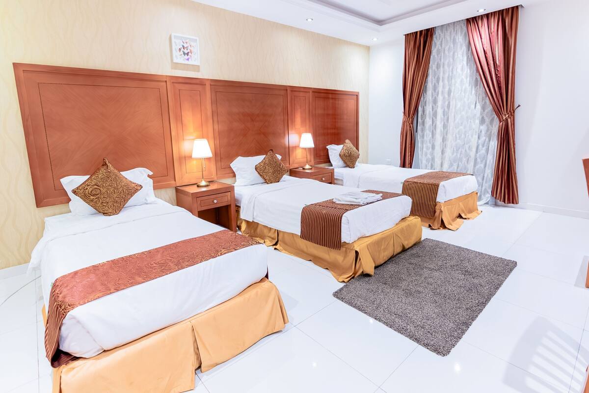 A hotel room with three beds, wooden headboard, white bedding, and a grey rug.