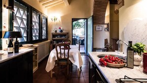 Une cuisine chaleureuse avec un coin repas, comprenant une table en bois, des chaises et un plan de travail sur lequel se trouve un saladier rempli de tomates.