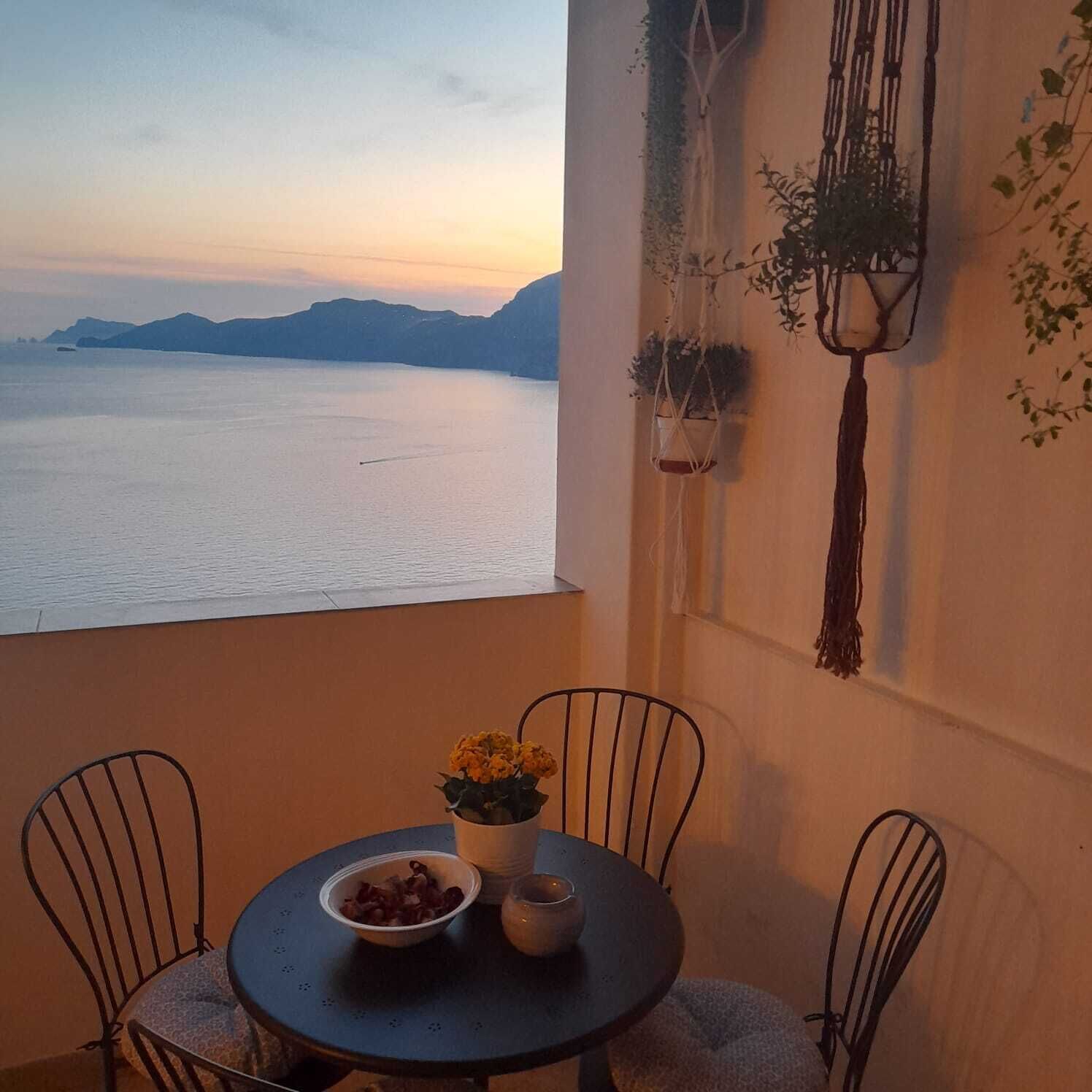 Ein Balkon mit Tisch, Stühlen und Blick auf das Meer bei Sonnenuntergang.