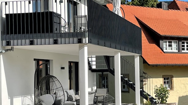 Une maison moderne à deux étages, dotée d’un balcon, d’un espace extérieur aménagé et d’un toit recouvert de tuiles rouges.