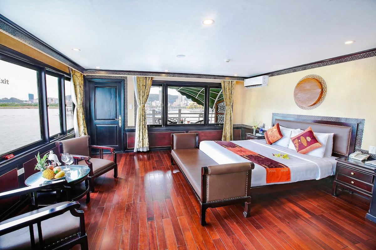 Une chambre d’hôtel avec un lit, une table avec des fruits et un verre, un canapé et une vue sur l’eau.