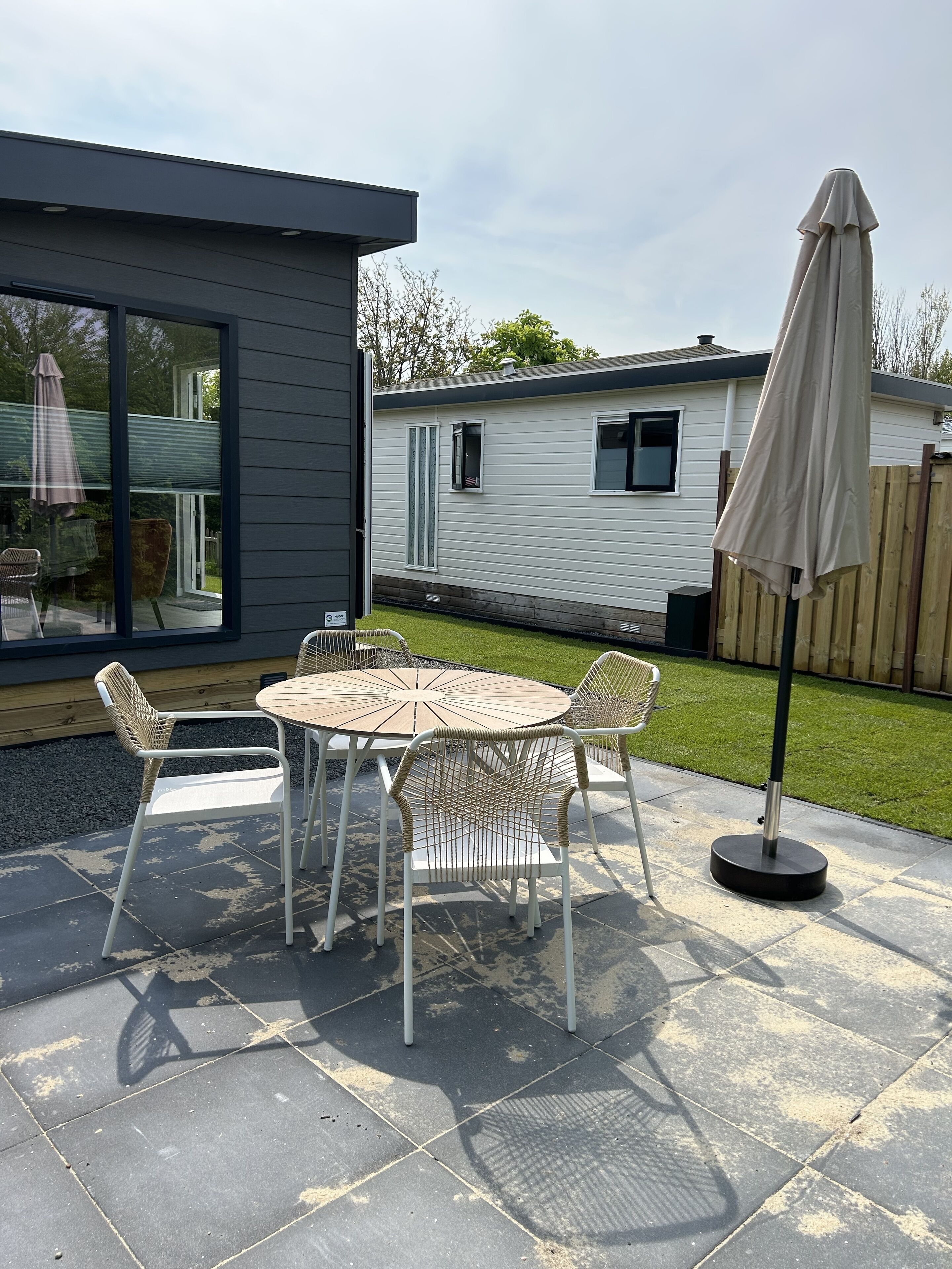 Un espace patio avec une table ronde et des chaises, un parasol et, au loin, une vue sur une maison mobile.