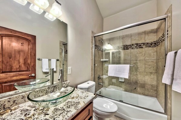 A modern bathroom with a glass shower enclosure, a glass sink, and a wooden door.