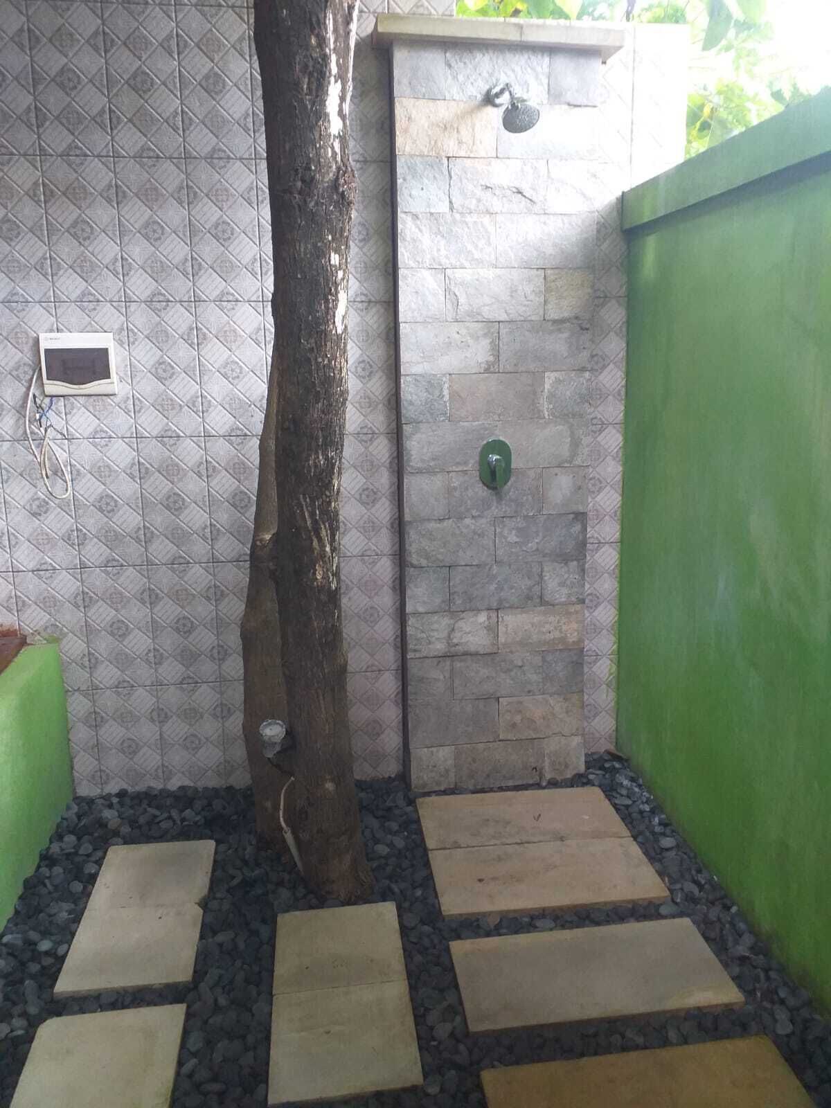 A small, enclosed outdoor shower area with a tiled wall, a wooden tree trunk, and a green wall.