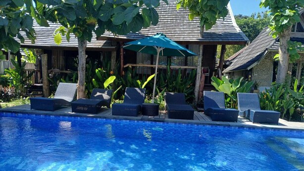 Una piscina con sillas de descanso y una sombrilla, rodeada de plantas tropicales y un edificio de madera.
