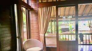 Una habitación con una silla de mimbre, una mesita con un control remoto y una puerta que da a un área exterior con terraza de madera y vegetación.