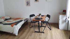 A small, neatly arranged room with a bed, a table with chairs, a small cabinet, and two framed pictures on the wall.