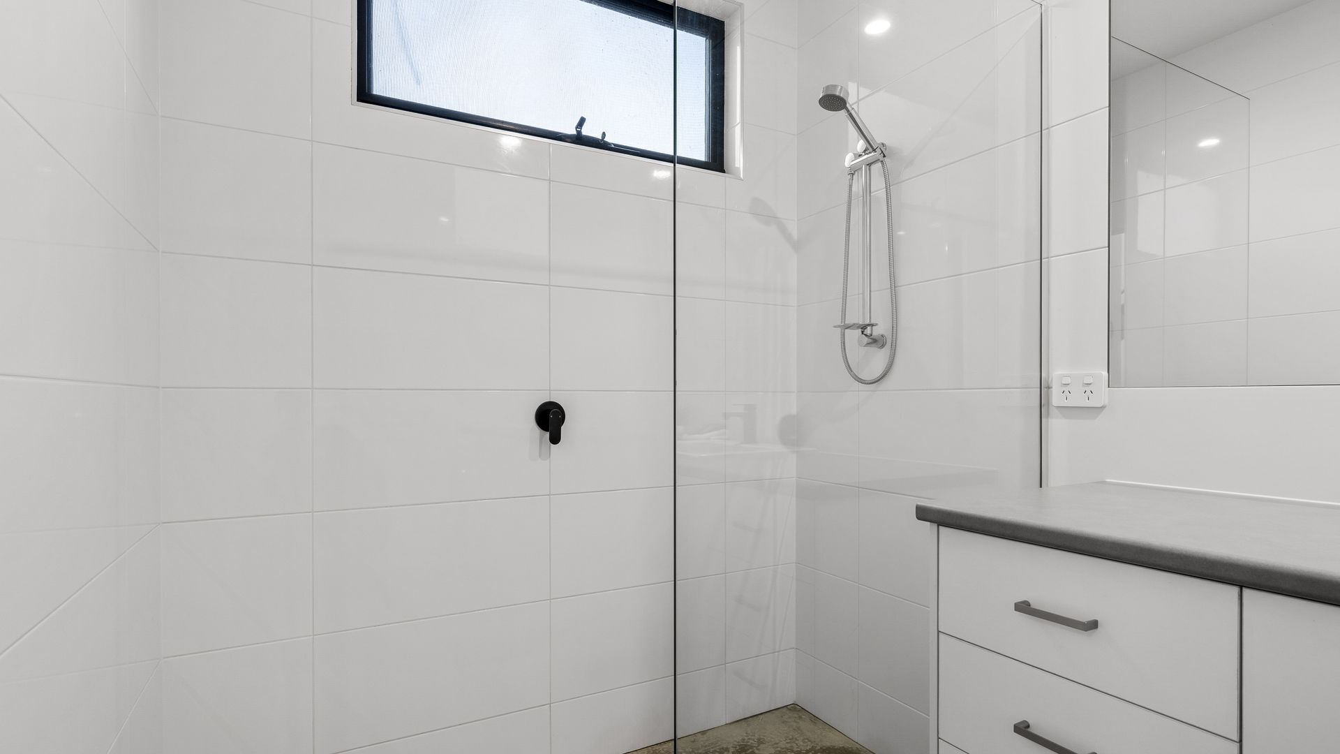 Un baño moderno con azulejos blancos, cabina de ducha de vidrio, ducha de mano y tocador con espejo y cajones.
