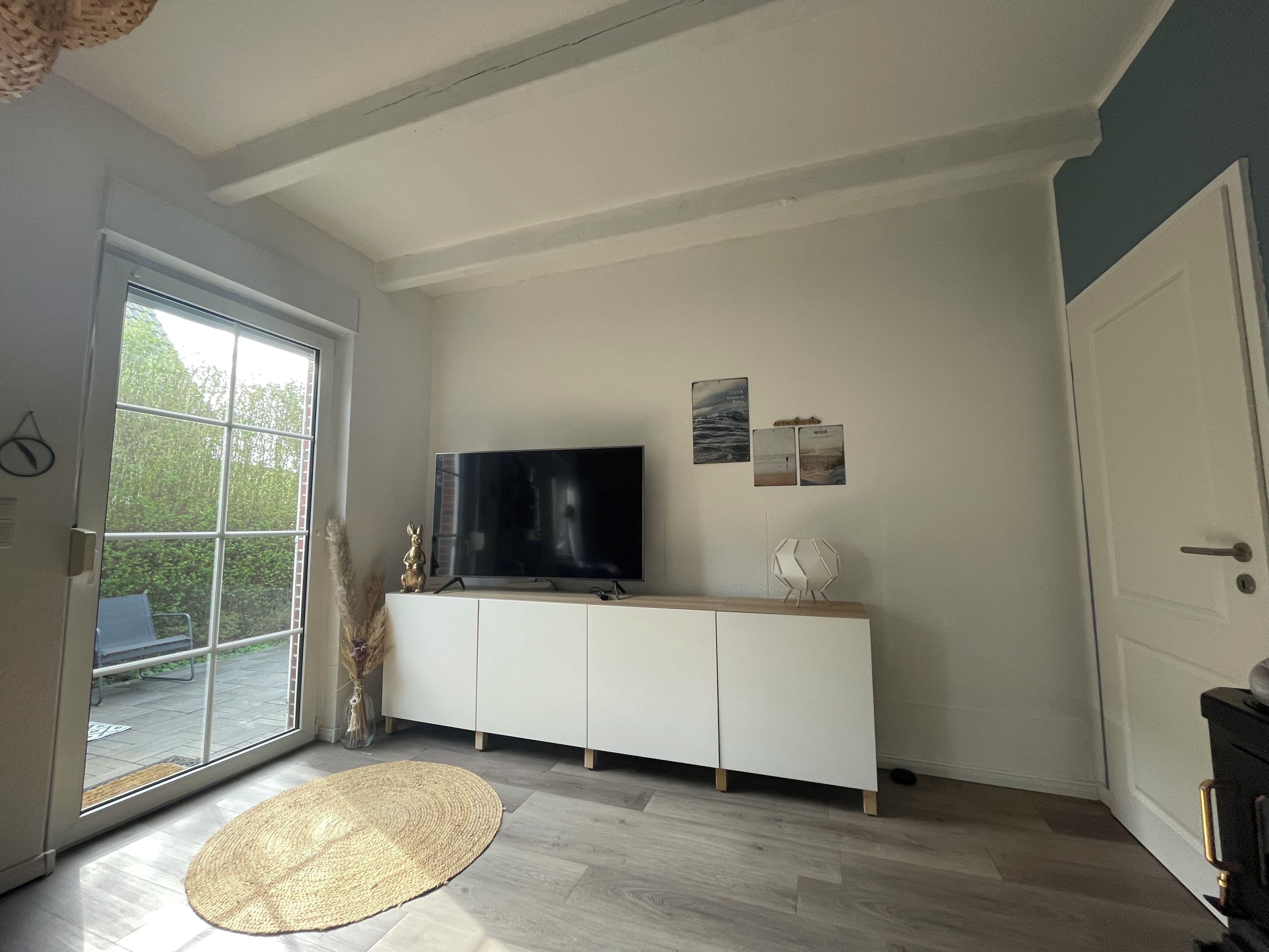 A room with a large window, a flat-screen TV on a white cabinet, a woven rug on the floor, and a door on the right.