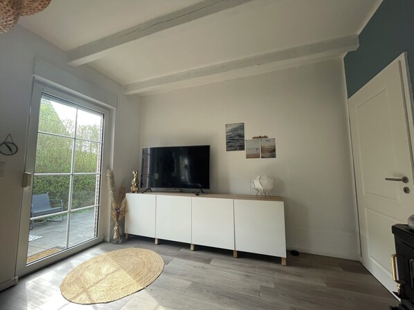 A room with a large window, a flat-screen TV on a white cabinet, a woven rug on the floor, and a door on the right.