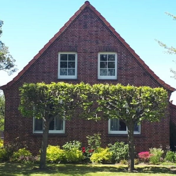A two-story brick house with a gabled roof, two windows, and a well-maintained garden with various plants and a tree.