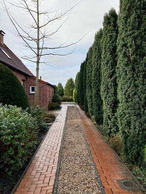 Un chemin pavé de briques mène à un bâtiment également construit en briques et surmonté d’un toit en tuiles, le tout étant entouré d’arbres et d’arbustes à feuilles persistantes.