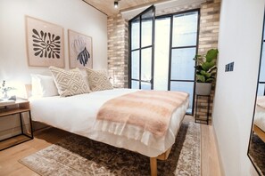 A bedroom with a large bed, two wall-mounted artworks, a potted plant, and a view through a large window.