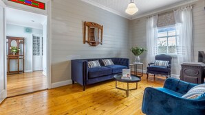 Un salón con un sofá azul, una mesa de centro redonda y un sillón azul. Se observa un aparador de madera con un espejo, una ventana con cortinas blancas y un piso de madera.