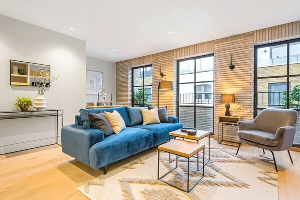 A modern living room with a blue sofa, a wooden accent wall, large windows, and a small side table with a lamp.