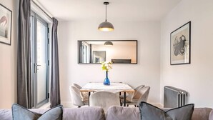 Ein modernes Wohnzimmer mit einer Couch, einem Esstisch mit Stühlen, einer Vase mit Blumen, einer Pendelleuchte, einem großen Spiegel und einer Heizung.