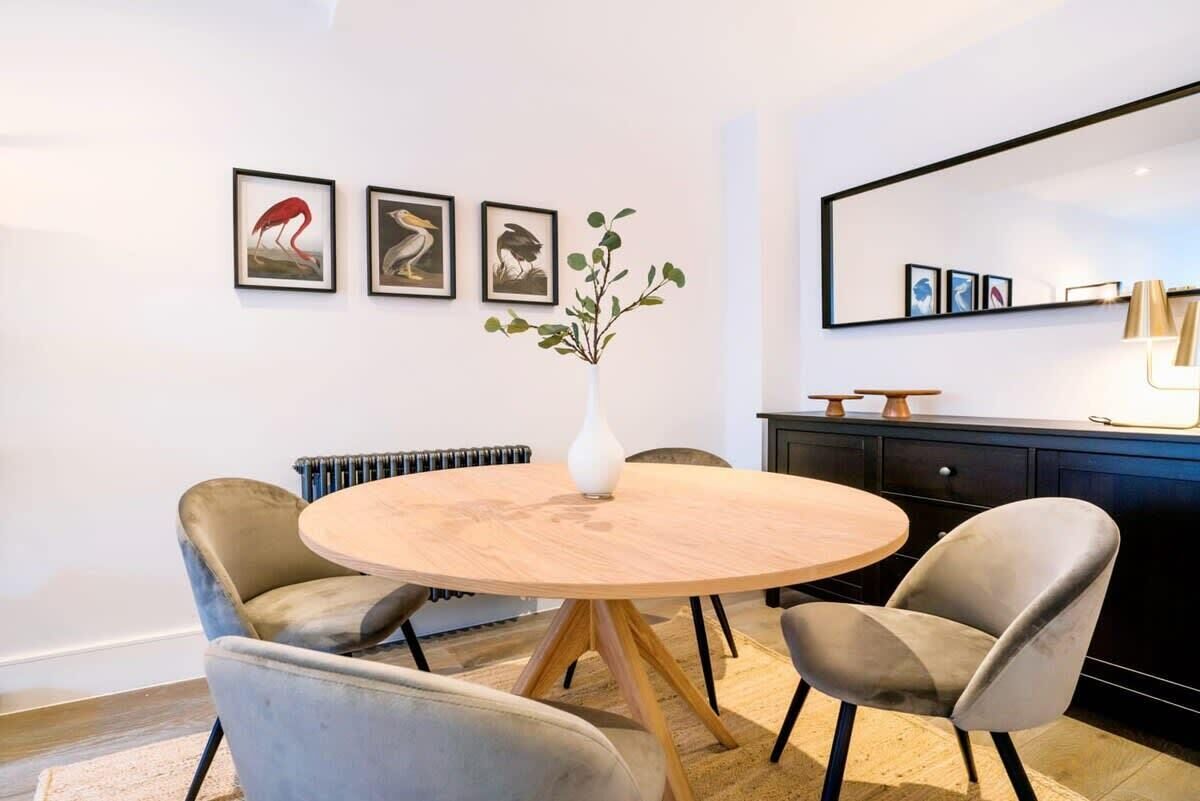 Ein stilvoller Essbereich mit einem runden Holztisch, vier grau gepolsterten Stühlen, einer weißen Vase mit Grünpflanzen und gerahmten Kunstwerken an der Wand.