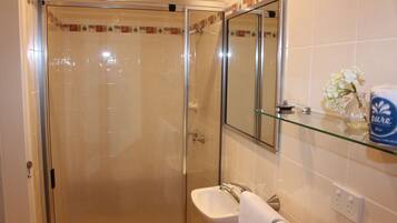A bathroom with a glass shower enclosure, a white sink, a mirror, and a glass shelf with toiletries.