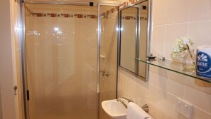 Une salle de bain avec une douche à parois de verre, un lavabo blanc, un miroir et une étagère en verre où sont rangés les articles de toilette.