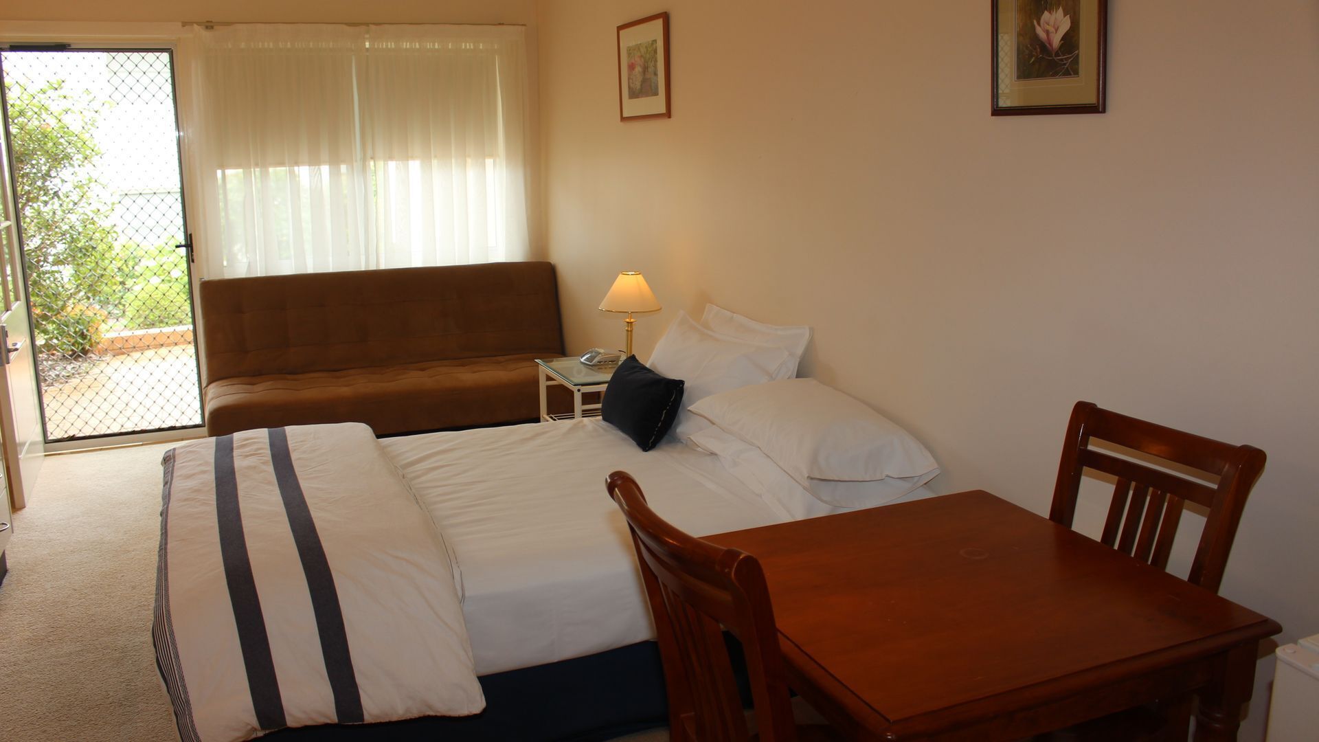 Habitació d'hotel amb un llit, una taula de fusta, una cadira, una petita taula de nit amb una làmpada i una vista per una porta de vidre.