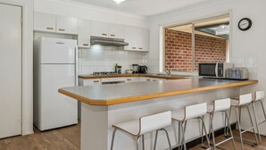 A modern kitchen with white cabinets, a stainless steel range hood, a built-in microwave, and a coffee maker. There are bar stools and a window with a brick wall view.