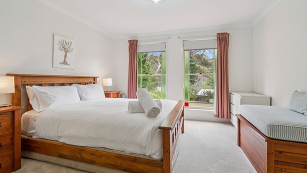 A bedroom with a wooden bed, a bedside table with a lamp, a window with curtains, and a view of greenery outside.