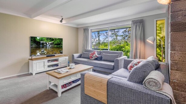Ein Wohnzimmer mit grauem Sofa, einem Couchtisch, einem Fernseher auf einem weißen Schrank und einem großen Fenster mit Vorhängen.