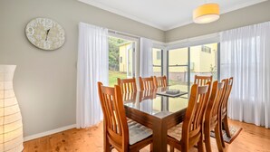 Un coin repas avec une table en verre entourée de chaises en bois, une horloge au mur et une vue sur un bâtiment jaune, visible à travers de grandes fenêtres.