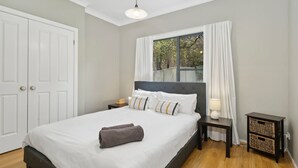 Un lit bien fait, avec du linge de lit blanc et une serviette grise, une table de chevet avec une lampe, une fenêtre avec des rideaux blancs et une porte.