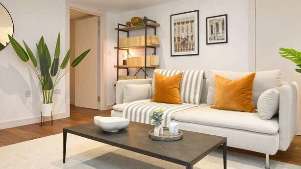 A modern living room with a white sofa, two orange cushions, a coffee table, a bookshelf, and a potted plant.