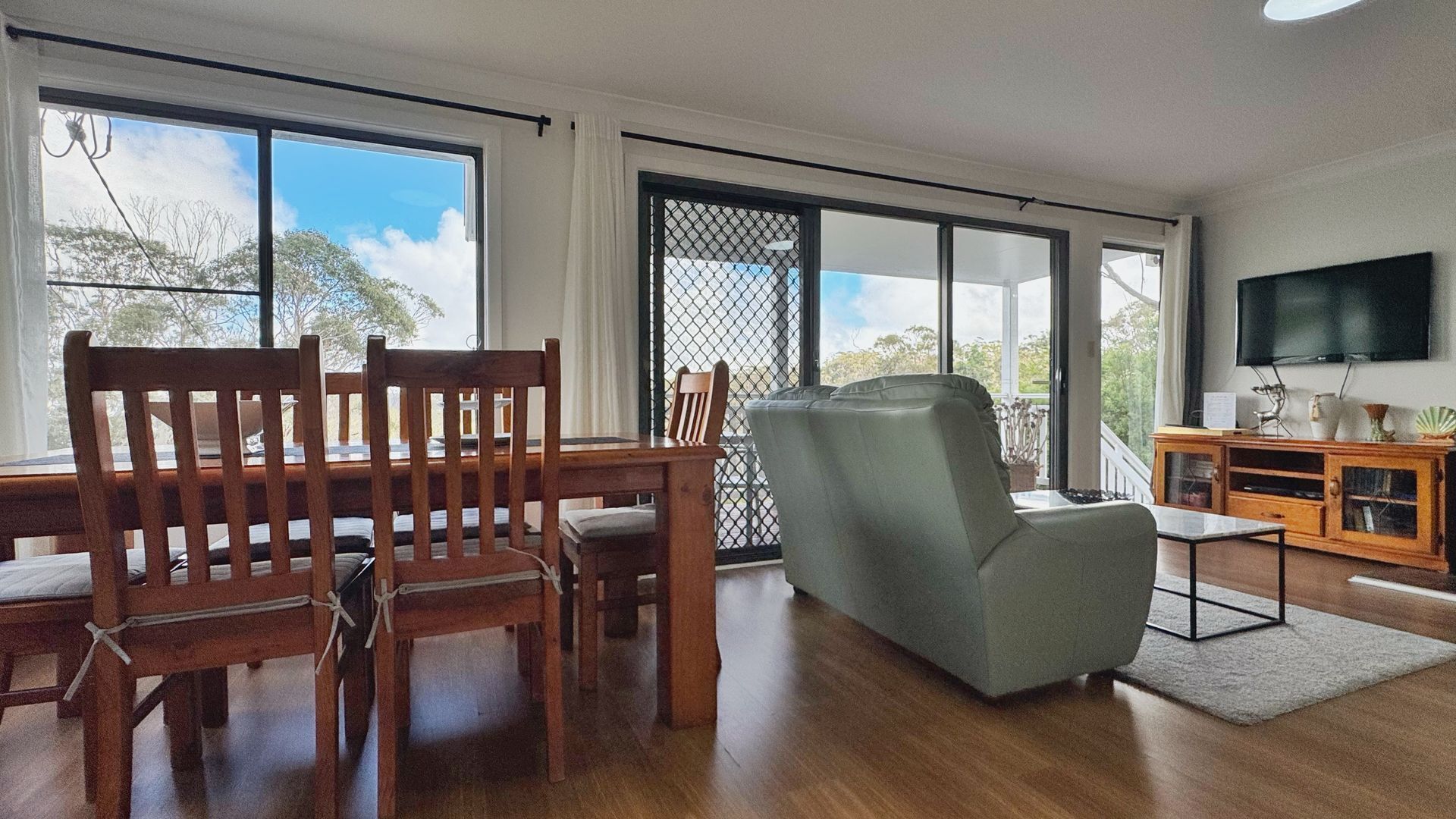 A spacious living area with a dining table, chairs, a sofa, a TV, and large windows offering a view of the outdoors.