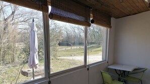 Een kamer met een tafel en twee stoelen, grote ramen met jaloezieën en uitzicht op een grasveld en bomen.